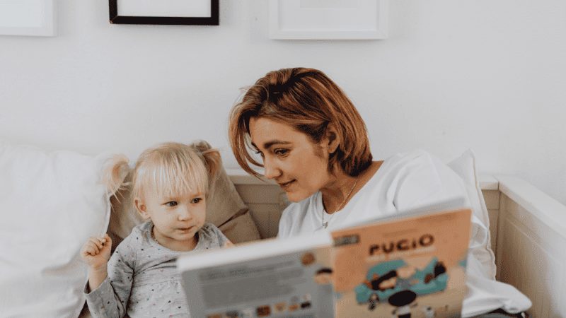 A parent and a toddler curled up in a cozy reading nook, with the child excitedly pointing at a colorful picture book, illustrating the concept of dialogic reading and shared focus.