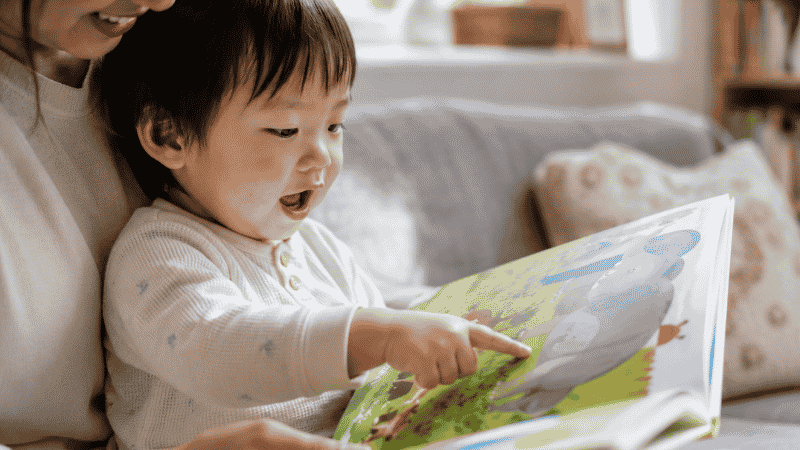 A toddler pointing at a colorful animal illustration in a picture book while a parent reads aloud, supporting language development