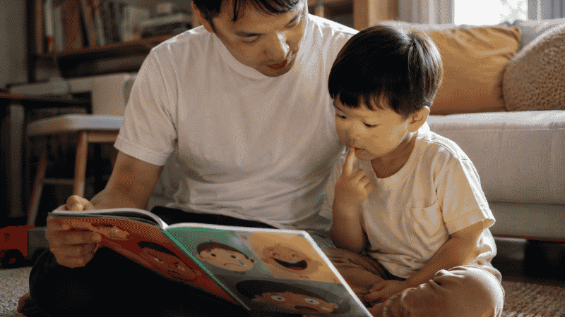 A parent and toddler sitting on the floor reading a colorful picture book about emotions, the child looking engaged and curious