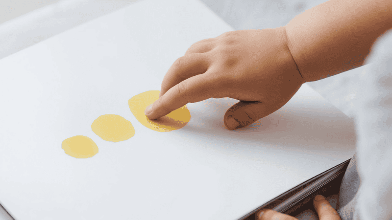 A close-up shot of a small child's hand pressing a painted yellow dot on a minimalist book page, demonstrating how interactive books engage high-energy toddlers.