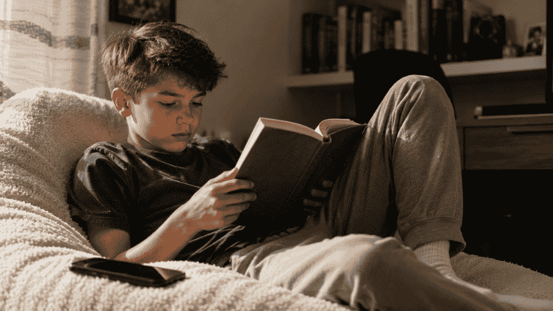 An 11-year-old child choosing to read a book instead of looking at their phone, sitting in a quiet corner absorbed in the story