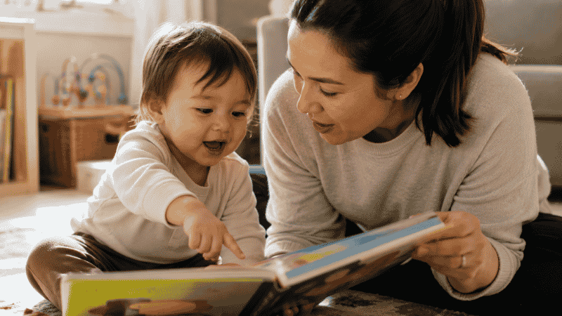 A toddler pointing at a picture in a board book while a parent listens and responds, practicing dialogic reading together