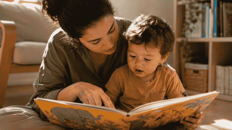 A parent reading a colorful picture book to a toddler, pausing to point at an illustration and name it together