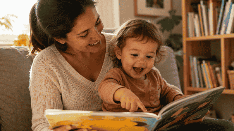 A 2-year-old toddler sitting with a parent reading a colorful picture book, the child clearly recognizing something in the story and reacting with delight